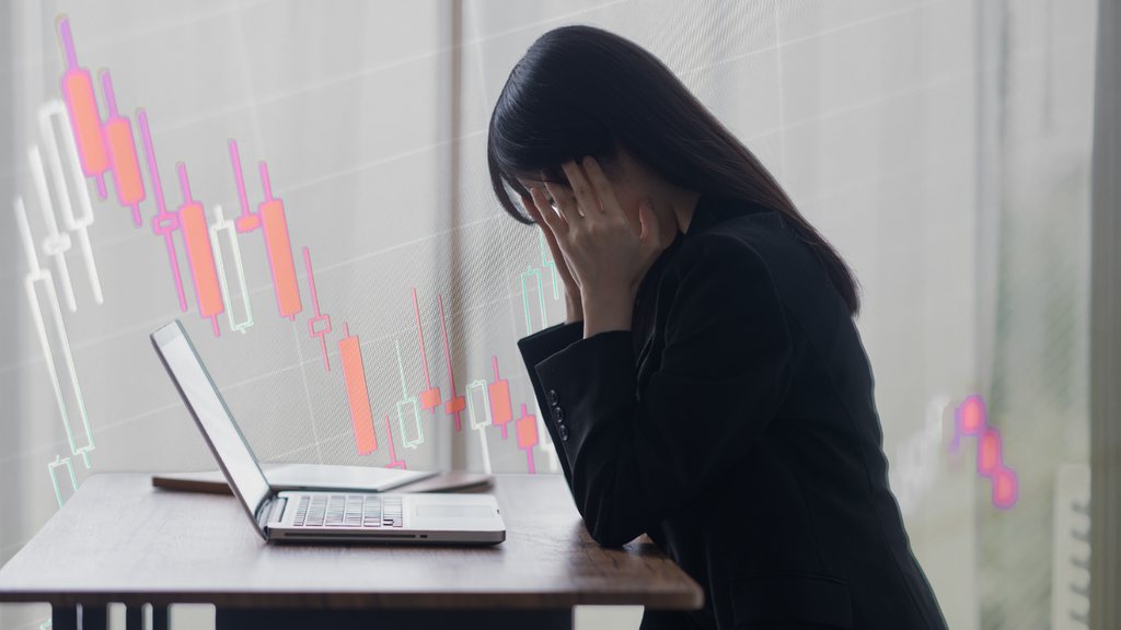 下落する株価チャートを背景に、パソコンの前で頭を抱える女性